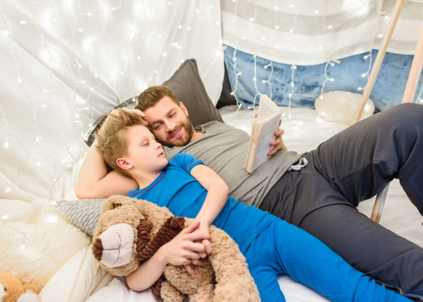 Happy father and son with teddy bear reading book in blanket fort